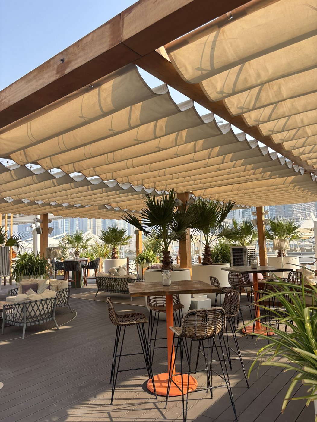 Ahşap iskeletli, bej kumaş gölgelikli bir kafe terası, küçük palmiyeler ve yüksek bar masalarıyla dolu