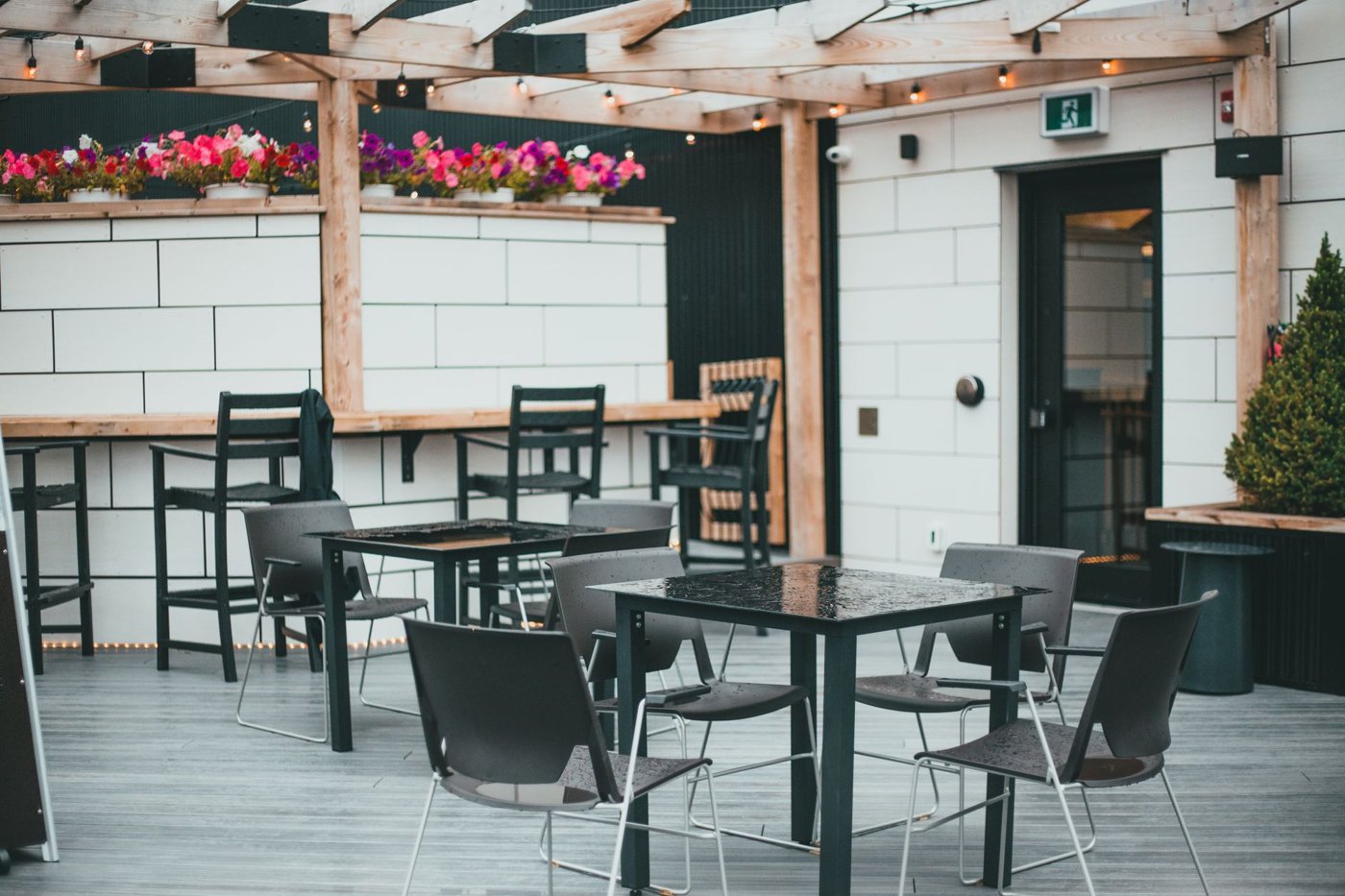 Ahşap pergolalı, beyaz blok duvarlı bir kafe terası, siyah kare masalar, sandalyeler ve renkli çiçekler içeriyor