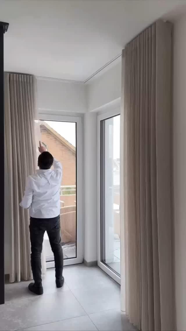 Beyaz gömlekli bir kişi, tavandan zemine uzanan açık bej renkli pileli fon perdeyi aralıyor