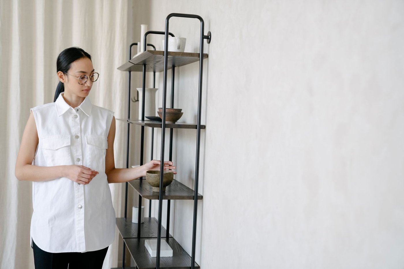 Siyah metal iskeletli ve koyu ahşap rafları olan minimalist bir ünitenin önünde duran kadın bir seramik kaseye uzanıyor