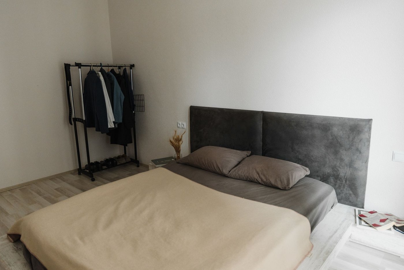 Koyu gri, kadife dokulu iki panelli yatak başlığına sahip, bej örtülü çift kişilik yatak ve minimalist oda